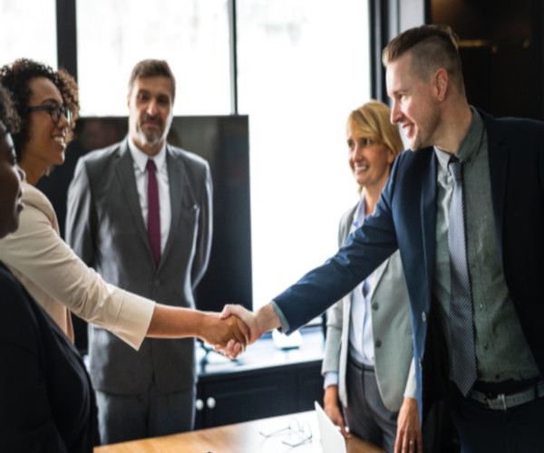 image of 5 people standing at table in office with 2 shaking hands across the table